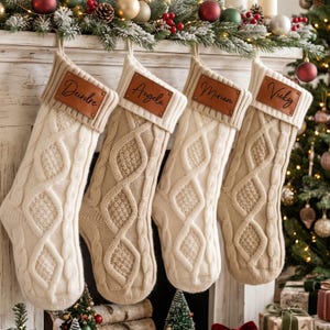 May include: Four cable-knit Christmas stockings in cream and beige tones, each featuring a ribbed cuff and a rectangular brown leather patch. The patches are engraved with the names Deirdre, Angela, Miriam, and Vicky in a cursive script font.