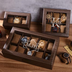 Puede incluir: Cajas de exhibici&oacute;n de relojes de madera con tapas de vidrio, que muestran varios relojes de pulsera. Las cajas son marrones con compartimentos individuales forrados con tela marr&oacute;n. Una caja tiene las iniciales "MJB" y otra el nombre "KEVIN" en el cristal.