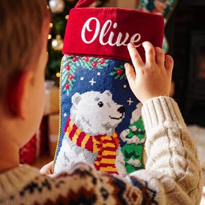 May include: A Christmas stocking featuring a needlepoint design of a white polar bear wearing a red and yellow striped scarf. The stocking has a red velvet cuff with the name Olive embroidered in white script and gold braided trim.