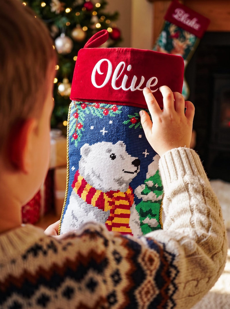 May include: A Christmas stocking featuring a needlepoint design of a white polar bear wearing a red and yellow striped scarf. The stocking has a red velvet cuff with the name Olive embroidered in white script and gold braided trim.