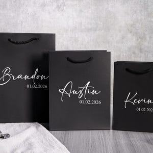 Puede incluir: Tres bolsas de regalo negras con nombres y fechas en escritura blanca. Las bolsas tienen asas de cuerda negra. Los nombres "Brandon", "Austin" y "Kevin" se muestran con la fecha "01.02.2026".