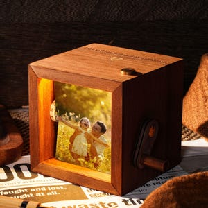 Peut inclure: Une boîte à musique en bois avec une photo de deux personnes. La boîte cubique est dotée d'une manivelle latérale et d'un petit bouton en laiton sur le dessus. La photo est éclairée de l'intérieur, créant une ambiance chaleureuse et nostalgique.