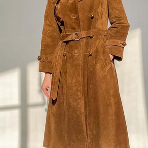 May include: A brown suede trench coat with a double-breasted design, a belt, and pockets. The coat has a classic collar and buttoned cuffs. The coat's length extends to the mid-calf. The coat is made of a soft, textured material.