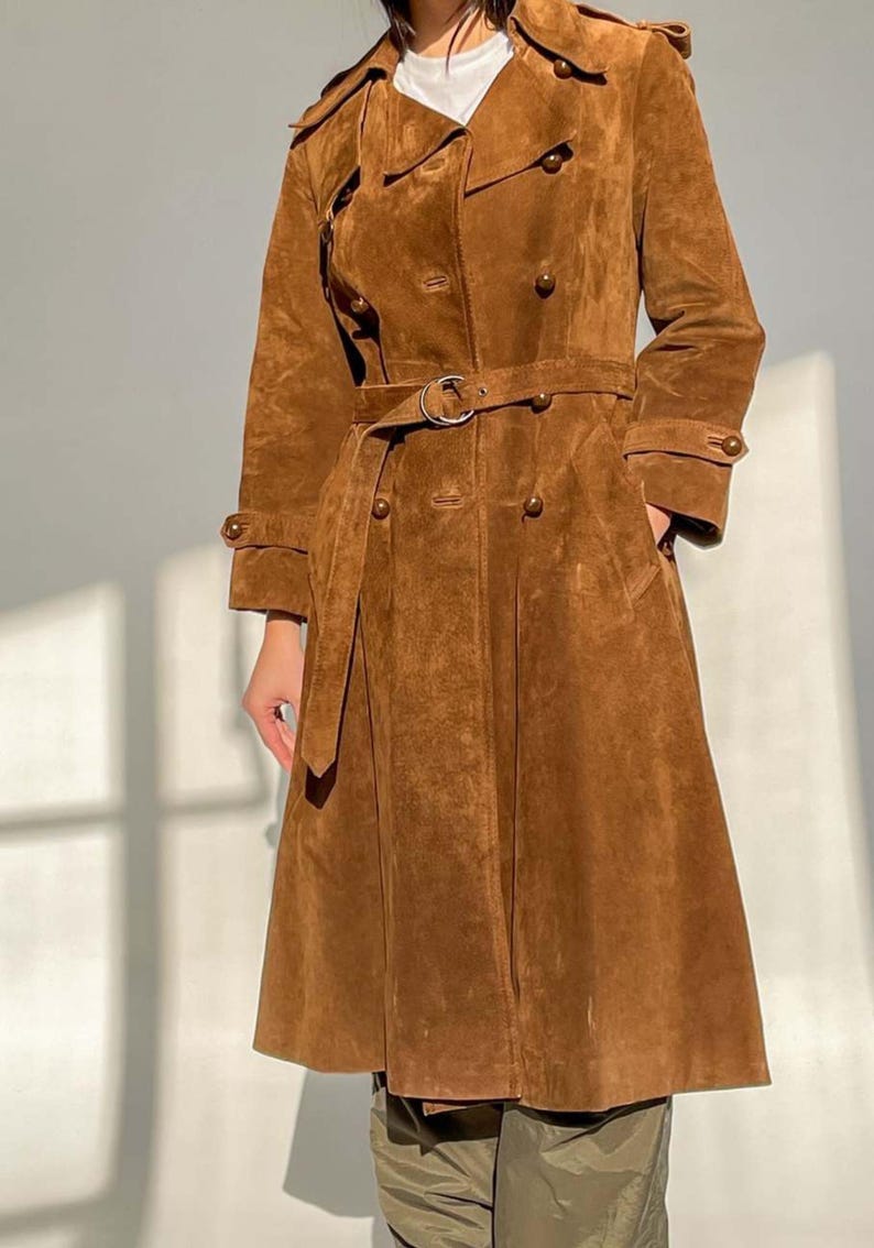 May include: A brown suede trench coat with a double-breasted design, a belt, and pockets. The coat has a classic collar and buttoned cuffs. The coat's length extends to the mid-calf. The coat is made of a soft, textured material.