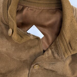 May include: Close-up of a brown suede jacket with a ribbed collar and button closure. The jacket has a satin lining and a classic design. The buttons are a matching brown color.