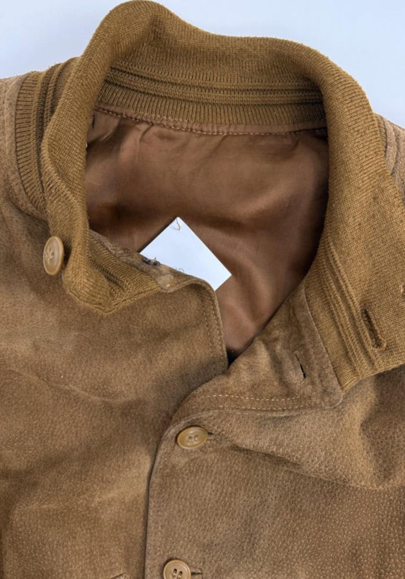 May include: Close-up of a brown suede jacket with a ribbed collar and button closure. The jacket has a satin lining and a classic design. The buttons are a matching brown color.