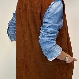 May include: A brown suede vest with a denim shirt underneath. The vest has a long, straight cut and a pocket on the side. The denim shirt has long sleeves. A gold watch is visible on the wrist.