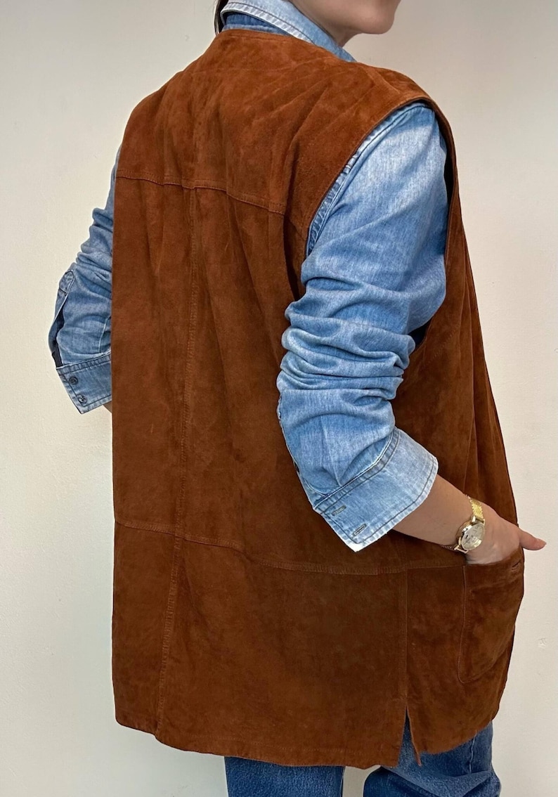 May include: A brown suede vest with a denim shirt underneath. The vest has a long, straight cut and a pocket on the side. The denim shirt has long sleeves. A gold watch is visible on the wrist.