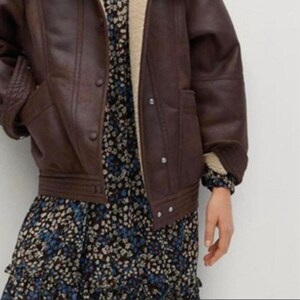 May include: A brown faux leather jacket with a shearling lining and button closures. The jacket is worn over a floral print dress with blue and white flowers.