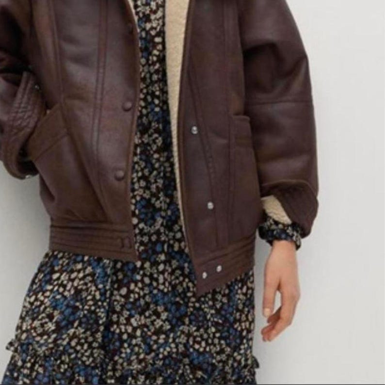 May include: A brown faux leather jacket with a shearling lining and button closures. The jacket is worn over a floral print dress with blue and white flowers.