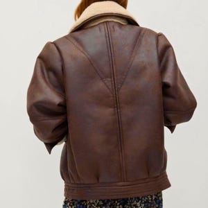 May include: A brown faux leather jacket with a cream-colored shearling collar. The jacket has a bomber-style design with a ribbed hem and cuffs. The back of the jacket features a V-shaped seam detail. The jacket is a medium brown color.