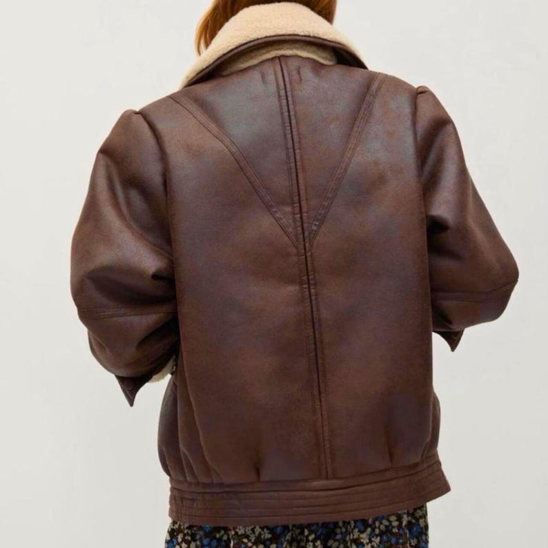 May include: A brown faux leather jacket with a cream-colored shearling collar. The jacket has a bomber-style design with a ribbed hem and cuffs. The back of the jacket features a V-shaped seam detail. The jacket is a medium brown color.
