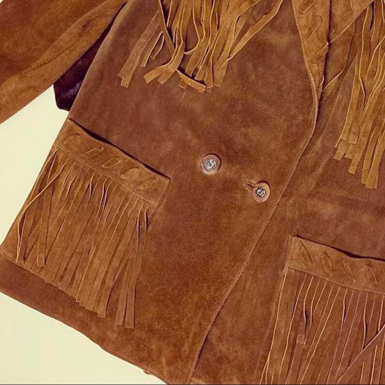 May include: Brown suede jacket with fringe detailing on the pockets, shoulders, and back. The jacket has two front pockets and two buttons. The jacket is a warm brown color.