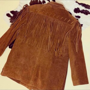 May include: A brown suede jacket with long sleeves and fringe detailing across the back and shoulders. The jacket is a medium brown color and has a classic Western style. The jacket is laid on a cowhide rug.