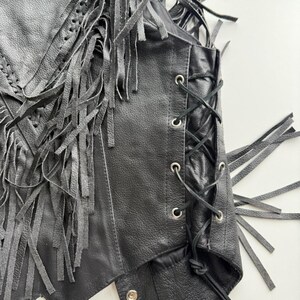 May include: Black leather vest with fringe detailing. The vest has a lace-up side and silver-tone eyelets. The front of the vest has a braided design. The vest is a classic biker style.