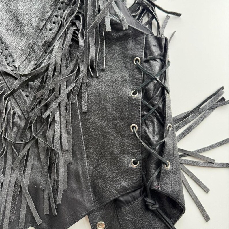 May include: Black leather vest with fringe detailing. The vest has a lace-up side and silver-tone eyelets. The front of the vest has a braided design. The vest is a classic biker style.