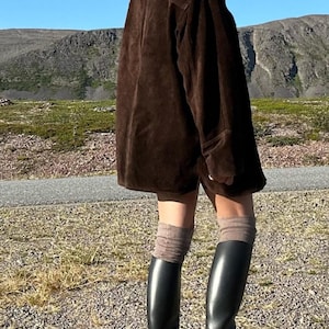 May include: A dark brown suede jacket with a collared neckline. The jacket is paired with black knee-high boots and brown socks. The background features a mountain range and a clear blue sky.