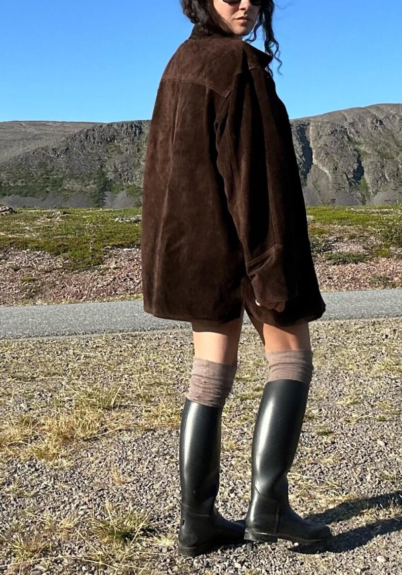 May include: A dark brown suede jacket with a collared neckline. The jacket is paired with black knee-high boots and brown socks. The background features a mountain range and a clear blue sky.