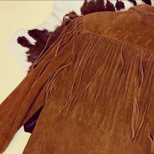 May include: A brown suede jacket with long fringe detailing on the back and sleeves. The jacket has a classic Western style, with a soft, textured appearance. The jacket is shown against a backdrop of a cowhide rug.