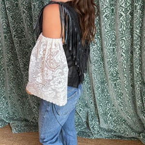 May include: A black leather vest with fringe detail and a white lace sleeve. The sleeve is made of a delicate floral lace pattern. The person is wearing blue jeans.