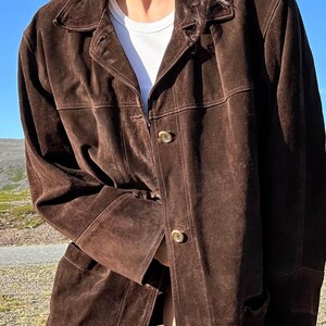 May include: A dark brown suede jacket with a collared neckline and button closure. The jacket has a classic design with visible stitching and two front pockets. The sleeves are long, and the jacket appears to be worn over a white t-shirt.