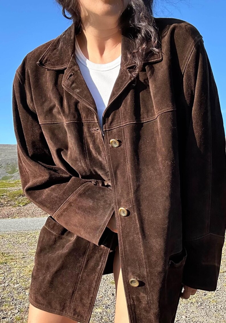 May include: A dark brown suede jacket with a collared neckline and button closure. The jacket has a classic design with visible stitching and two front pockets. The sleeves are long, and the jacket appears to be worn over a white t-shirt.