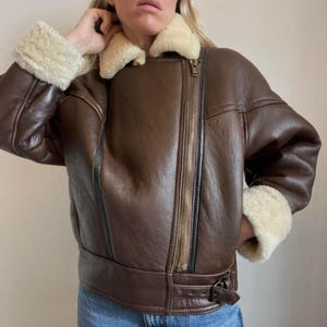 May include: A brown leather jacket with a shearling collar and cuffs. The jacket features a zipper closure and a belt at the waist. The shearling is a cream color. The jacket is a classic style.