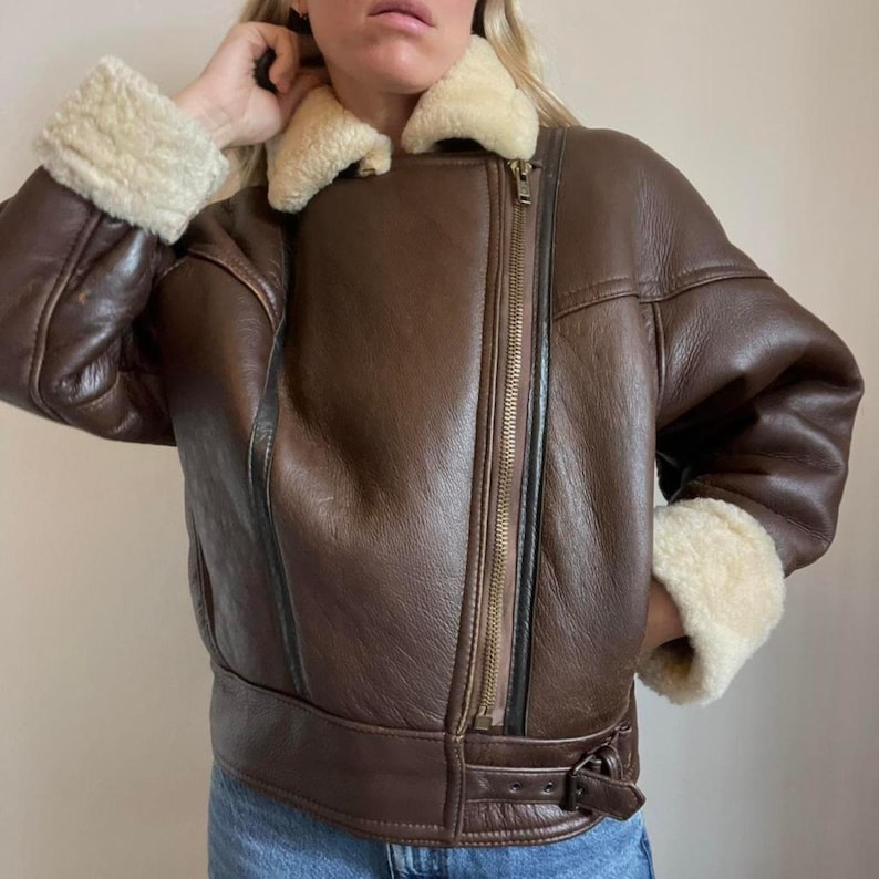 May include: A brown leather jacket with a shearling collar and cuffs. The jacket features a zipper closure and a belt at the waist. The shearling is a cream color. The jacket is a classic style.