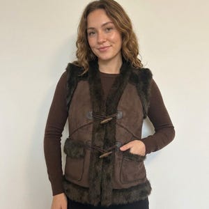 May include: A brown suede vest with dark brown faux fur trim and wooden toggle fastenings. The vest features two front pockets and is worn over a brown long-sleeved top. A stylish outerwear garment.