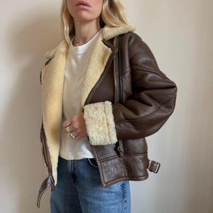 May include: A brown leather jacket with a cream-colored shearling lining and collar. The jacket features a zip closure, a buckled belt detail, and shearling cuffs. The jacket is a classic style, perfect for cold weather.