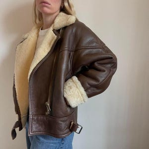 May include: A brown leather jacket with a cream-colored shearling lining and trim. The jacket features a zip closure, a buckled belt detail, and shearling cuffs. The jacket is a classic style, suitable for cold weather.
