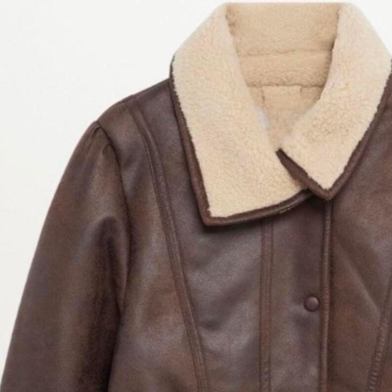 May include: A close-up of a brown faux leather jacket with a cream-colored shearling collar. The jacket has a button closure and a classic design, perfect for cold weather. The jacket's measurements are not visible.