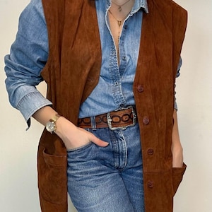 May include: A brown suede vest with large pockets and button closures. The vest is worn over a denim shirt and paired with blue jeans. A brown leather belt with a silver buckle and a gold watch complete the look. A vintage-inspired outfit.