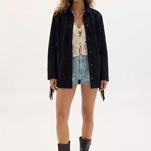 May include: A black suede jacket with fringe detailing, worn over a white top and denim shorts. The outfit is completed with black leather boots. The jacket has fringe on the chest, sleeves, and back.
