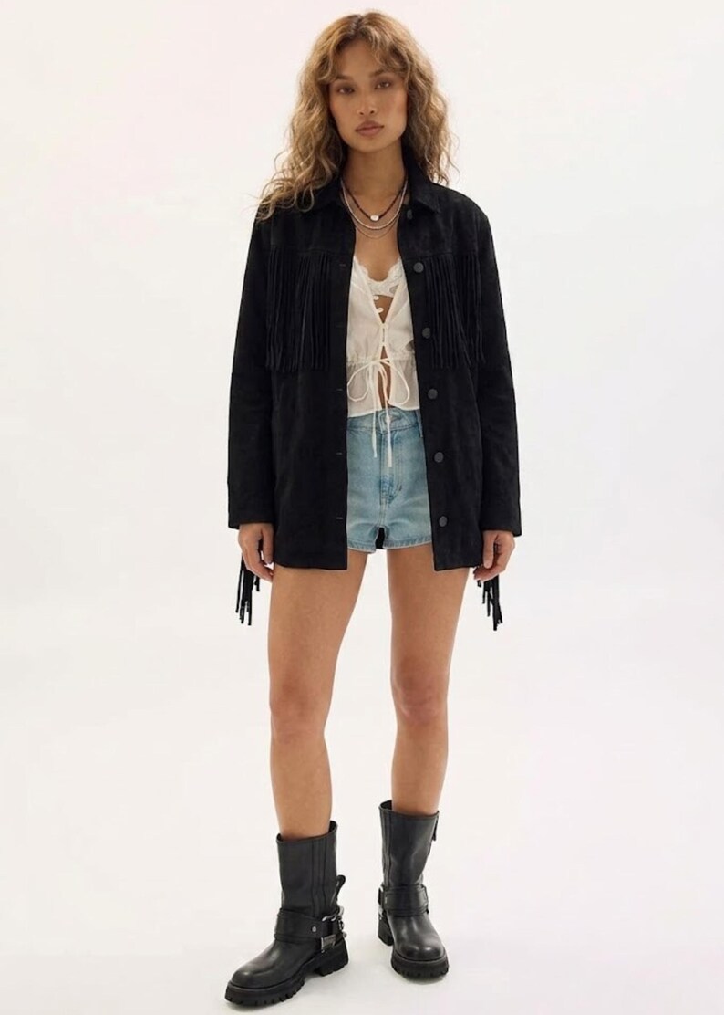May include: A black suede jacket with fringe detailing, worn over a white top and denim shorts. The outfit is completed with black leather boots. The jacket has fringe on the chest, sleeves, and back.