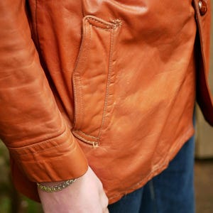 May include: Close-up of a vintage, burnt orange leather jacket. The jacket features a tailored fit, a front pocket, and a belt detail. The leather shows signs of wear, adding to its character. The sleeve is visible.