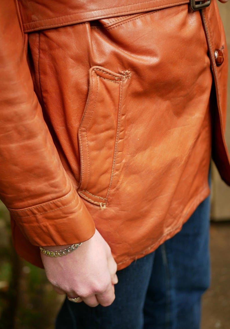 May include: Close-up of a vintage, burnt orange leather jacket. The jacket features a tailored fit, a front pocket, and a belt detail. The leather shows signs of wear, adding to its character. The sleeve is visible.