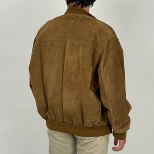 May include: A brown suede bomber jacket with a ribbed hem and cuffs. The jacket has a classic design with a collar and a front zipper. The model is wearing khaki pants. The jacket is a versatile piece for any wardrobe.
