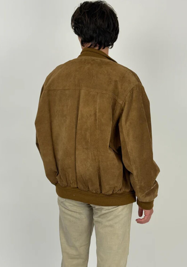 May include: A brown suede bomber jacket with a ribbed hem and cuffs. The jacket has a classic design with a collar and a front zipper. The model is wearing khaki pants. The jacket is a versatile piece for any wardrobe.