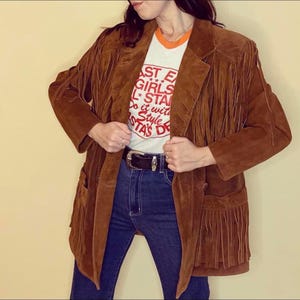 May include: A brown suede jacket with fringe detailing. The jacket has a classic blazer cut with pockets and a button closure. The model is wearing a white t-shirt with red text and blue jeans. A black belt with a silver buckle is visible.