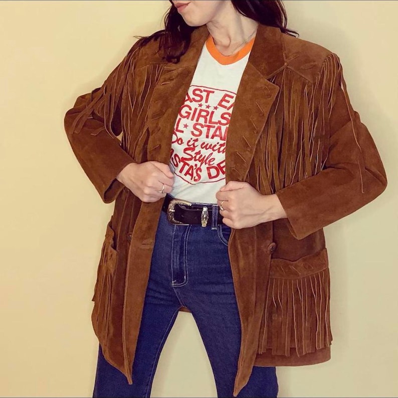 May include: A brown suede jacket with fringe detailing. The jacket has a classic blazer cut with pockets and a button closure. The model is wearing a white t-shirt with red text and blue jeans. A black belt with a silver buckle is visible.