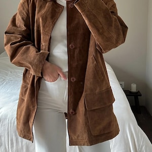 Könnte beinhalten: Braune Wildlederjacke mit Kragen, Knopfverschluss und zwei Vordertaschen. Die Jacke wird über einem weißen Oberteil und einer weißen Hose getragen. Die Jacke hat eine lockere Passform und ein klassisches Design.