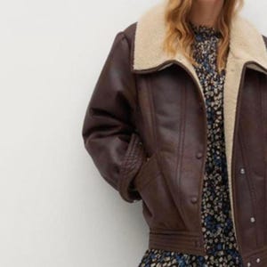 May include: A brown faux leather jacket with a cream-colored shearling collar. The jacket has a button-up front, a belt at the waist, and pockets. The sleeves have a quilted design. The jacket is a stylish outerwear piece.