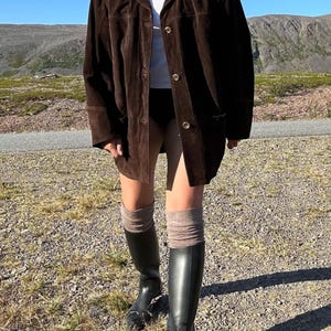 May include: A dark brown suede jacket with button closures. The jacket is worn over a white top and black shorts. The outfit is completed with knee-high black boots and tan socks. The background features a gravel path and a mountain range.