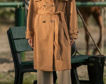 Puede incluir: Un abrigo tipo trench de color marrón claro con diseño de doble botonadura, cinturón a juego y detalles de botones. El abrigo se combina con pantalones color beige y botines marrones. El fondo incluye un banco verde y un caballo.