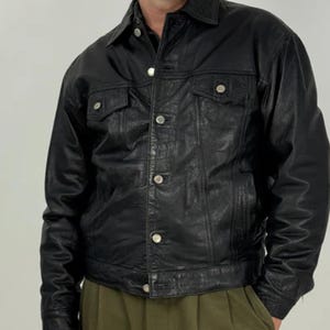 May include: Black leather jacket featuring a traditional collar, two chest pockets, and silver button fastenings. The jacket has a slightly aged look, possibly vintage. A classic piece of outerwear, suitable for various styles.