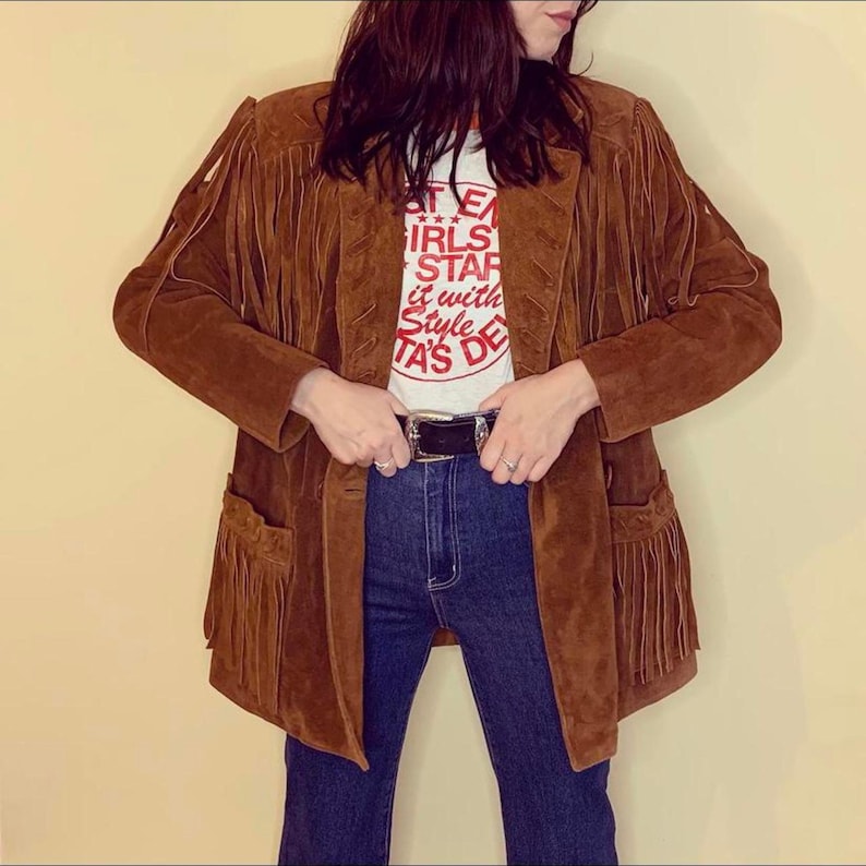 May include: Brown suede jacket with fringe detailing on the sleeves, pockets, and hem. The jacket is worn over a white graphic tee and blue jeans. A black belt with a silver buckle is visible.