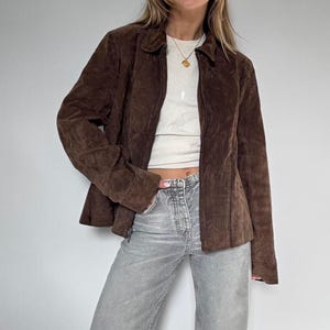 Peut inclure: Veste en daim marron avec un col et une fermeture éclair. La veste a des manches longues et une coupe ajustée. Le modèle porte un haut blanc et un jean gris. La veste semble être de style vintage ou rétro.