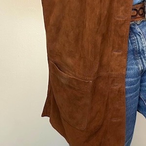May include: A brown suede vest with a single pocket and button closure. The vest is a warm, earthy tone and appears to be vintage. The garment is shown against a light background and denim jeans.