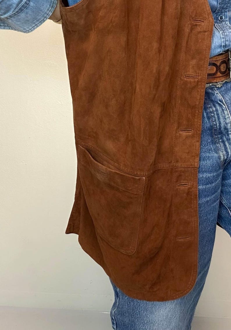 May include: A brown suede vest with a single pocket and button closure. The vest is a warm, earthy tone and appears to be vintage. The garment is shown against a light background and denim jeans.